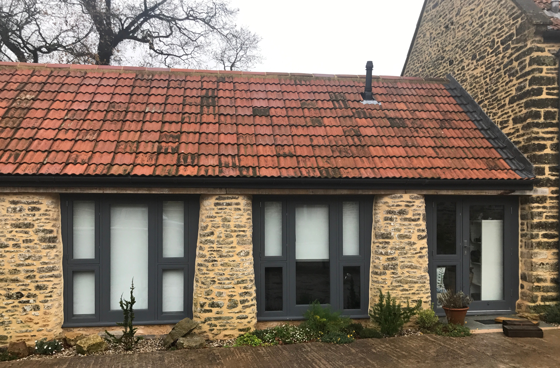Restored single-storey wing with reclaimed clay pantile roof and slim grey-framed windows set into original stone openings